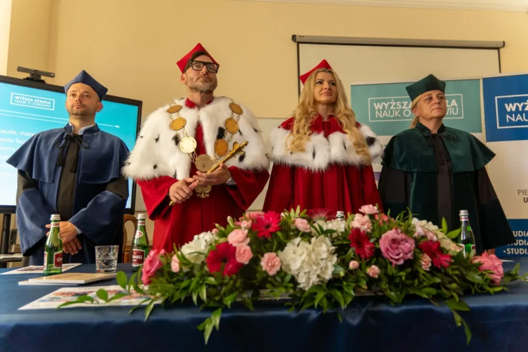 Na zdjęciu widoczni są rektor uczelni trzymający berło, prorektor oraz dziekani stojący za stołem prezydialnym w strojach akademickich podczas uroczystości dyplomatorium