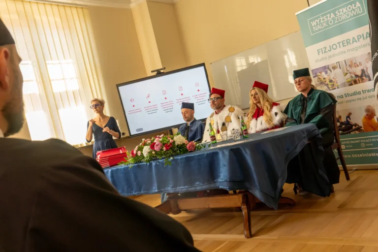 Na zdjęciu widoczna jest sala, w której za długim stołem prezydialnym, przykrytym obrusem, siedzą rektor uczelni, prorektor i dziekani.